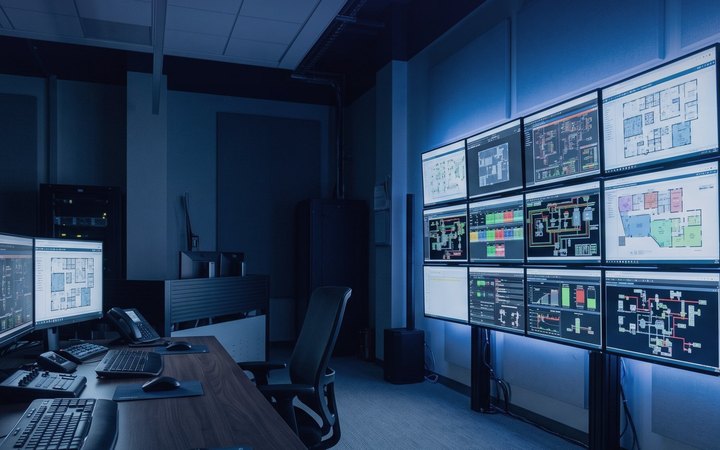 A building management control room at night: a video wall of BMS floor plans and schematics beside an operator desk.