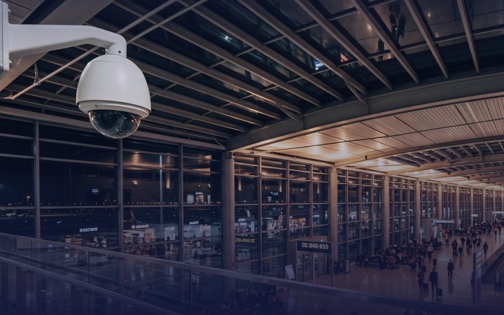A white dome CCTV camera on a wall bracket overlooking a long, busy airport terminal concourse.