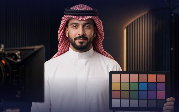 Illustrative studio scene: a presenter holds an uncalibrated 24-patch colour test chart beside a cinema camera.