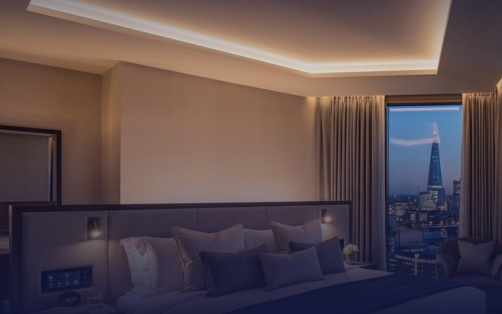 Hotel guest room at dusk with cove lighting, a bedside room control panel and a city skyline beyond the window.