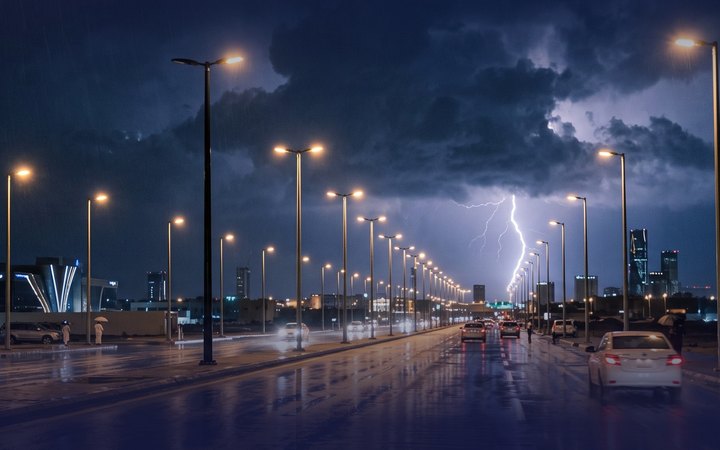 Lightning strikes beyond a rain-soaked city road lined with lit street lights on a stormy night.