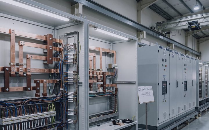 Two open low-voltage switchgear sections with copper busbars and wiring, beside a closed grey line-up on a workshop floor.